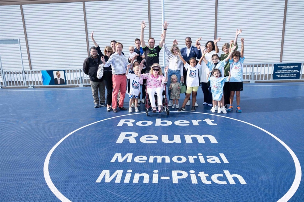 Courtney poses for a photo on the dark blue mini-pitch with his family members, coaches, and other community members. the text on the center circle of the pitch reads "robert reum memorial mini-pitch"